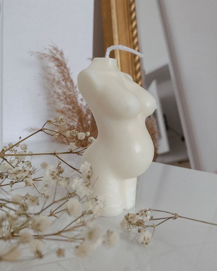 Vela Aromática Grávida Vela formato torso feminino grávido creme sobre mesa branca com flores secas e espelho