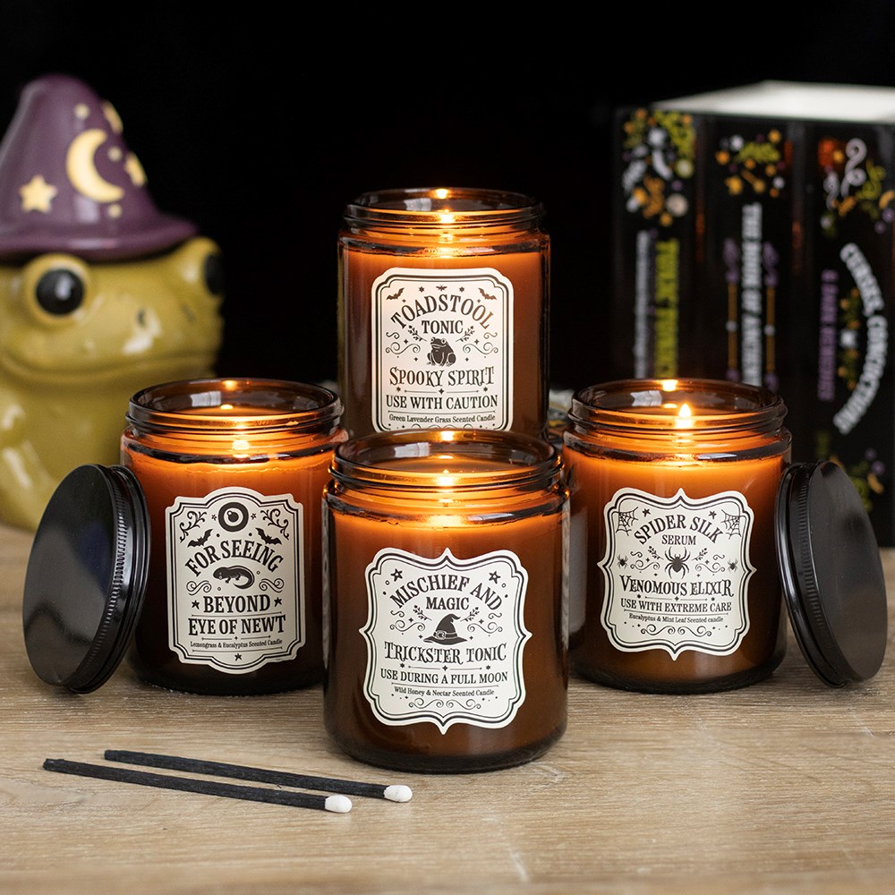 Quatro velas aromáticas em frascos de vidro âmbar com rótulos temáticos e um objeto decorativo de sapo ao fundo