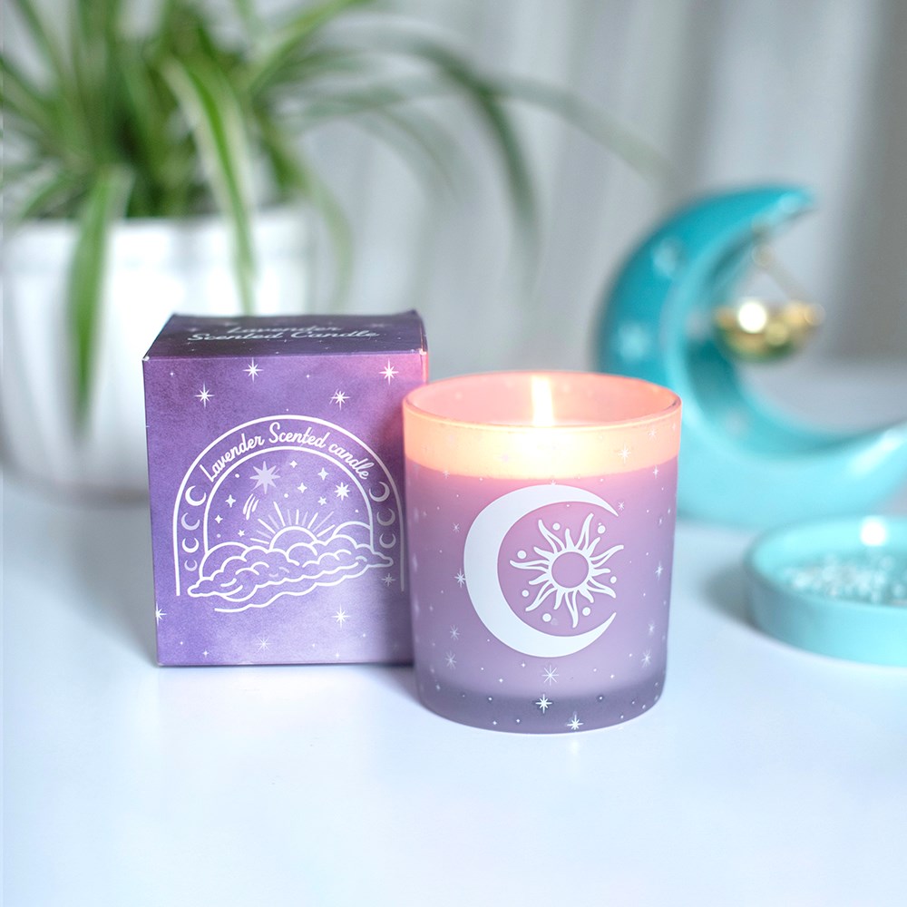 Vela aromática lilás com decoração de lua e sol junto a caixa roxa com ilustração similar, em mesa branca