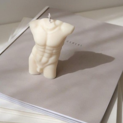 Vela escultórica branca em forma de torso masculino sobre revistas cinza