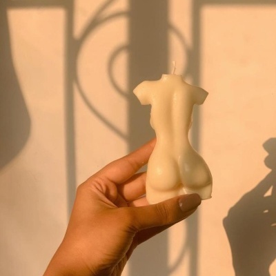 Vela creme em forma de corpo feminino segurada pela mão com luz natural ao fundo