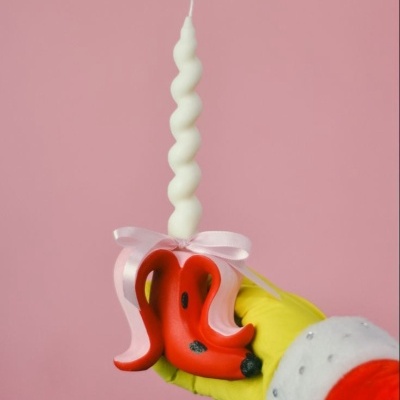 Vela decorativa branca espiral com base vermelha em formato orgânico, mão com luva verde e manga vermelha com punho branco segurando vela