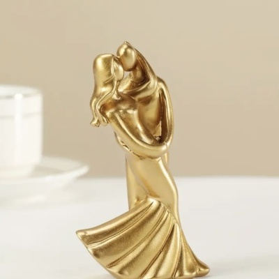 Escultura dourada de um casal em abraço numa superfície branca com fundo bege e chávena