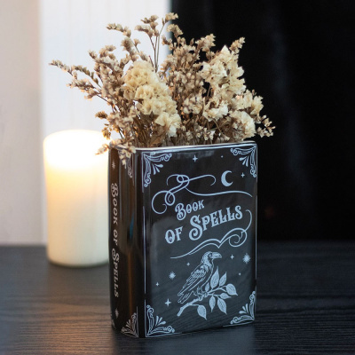 Vaso em forma de livro preto com texto e ilustração brancos, flores secas e vela acesa ao fundo