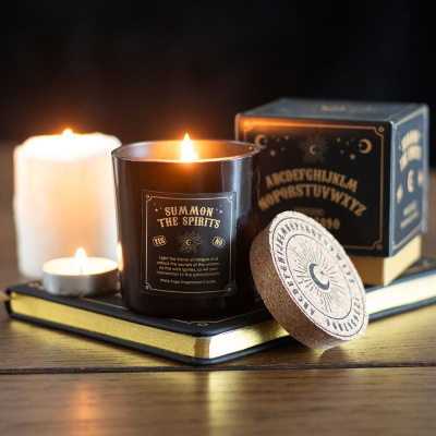 Vela aromática em copo preto com rótulo dourado sobre um caderno e caixa de vela com tampa de cortiça