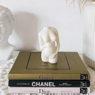 Vela em forma de torso feminino sobre livros de moda empilhados com uma estátua e uma jarra brancas ao redor.