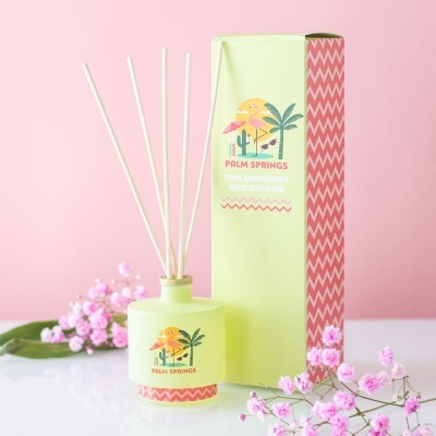 Difusor de aromas Palm Springs com embalagem verde e detalhes cor-de-rosa, flores e fundo rosa.