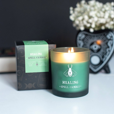 Vela verde com texto HEALING SPELL CANDLE e caixa preta com etiqueta verde com o mesmo texto em superfície branca