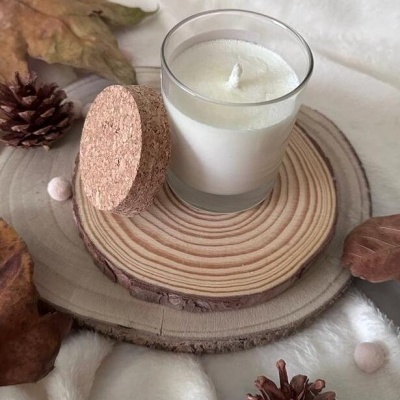 Vela branca em copo de vidro com tampa de cortiça sobre madeira, com decoração natural ao redor.