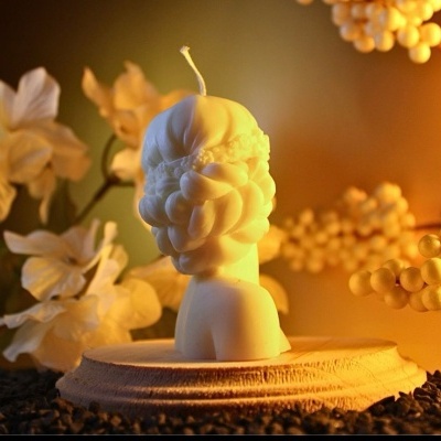 Vela branca decorativa em forma de cabeça com flores, em suporte redondo, com grãos de café e decoração desfocada ao fundo.