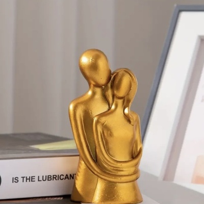 Escultura dourada de duas figuras humanas abraçadas, sobre mesa com livro e moldura