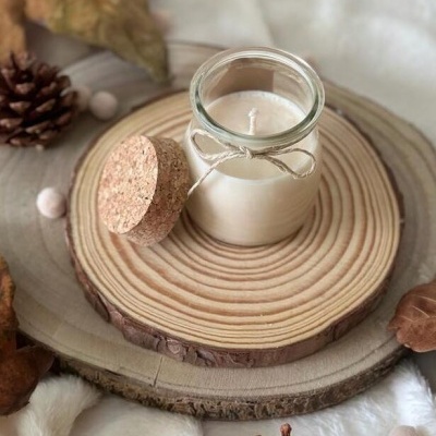 Vela branca em frasco de vidro com tampa de cortiça e cordão, sobre suportes de madeira e elementos naturais ao redor