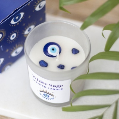 Vela perfumada White Sage com decoração de olho grego e pedras azuis, caixa azul ao fundo e folhas verdes ao lado.