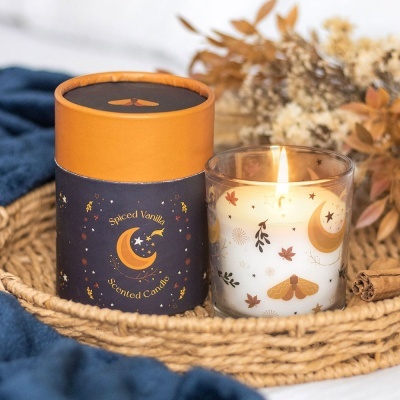 Vela aromática Spiced Vanilla com decoração de estrelas e lua, ao lado de embalagem cilíndrica, numa bandeja de palha com fundo azul e raminhos secos.