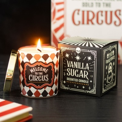 Vela acesa em vidro decorado com padrão de losangos castanhos e brancos, texto WELCOME TO THE CIRCUS e caixa preta com texto VANILLA SUGAR SCENTED CANDLE ao lado.