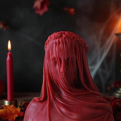 Vela decorativa vermelha em forma de cabeça coberta por véu com vela acesa e abóboras ao fundo