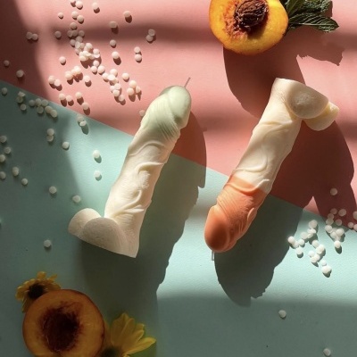 Duas velas em forma de pénis com pêssegos, flores amarelas e bolinhas brancas sobre fundo rosa e azul