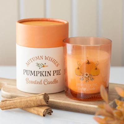 Vela perfumada Pumpkin Pie com embalagem laranja e branca e paus de canela sobre tábua de madeira.