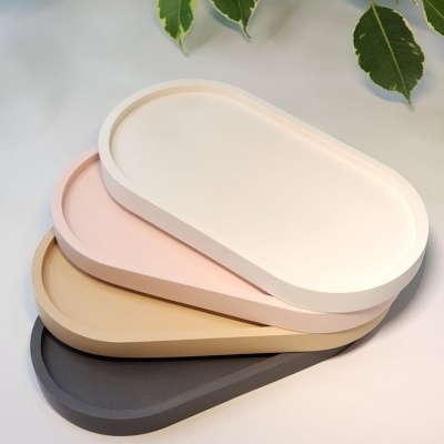 Conjunto de quatro tabuleiros ovais empilhados em cores pastel e cinza, sobre superfície branca com plantas ao fundo.