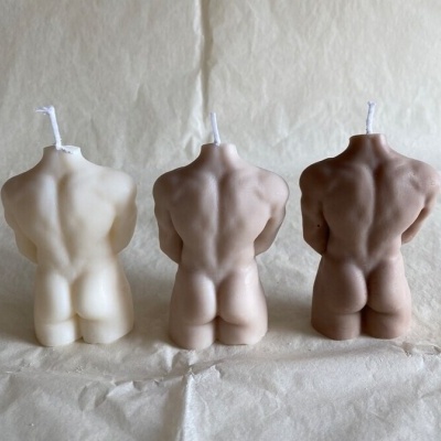 Velas decorativas em forma de torso masculino em três tons de pele diferentes, sobre tecido claro.