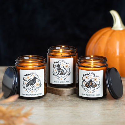 Três velas perfumadas em frascos de vidro âmbar com etiquetas ilustradas e uma abóbora laranja ao fundo
