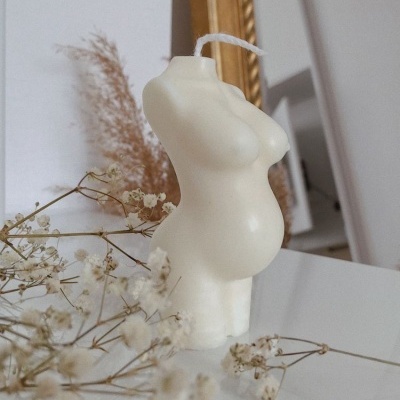 Vela formato torso feminino grávido creme sobre mesa branca com flores secas e espelho