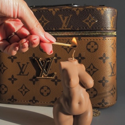 Vela em forma de tronco feminino acesa com fósforo junto a bolsa Louis Vuitton marrom