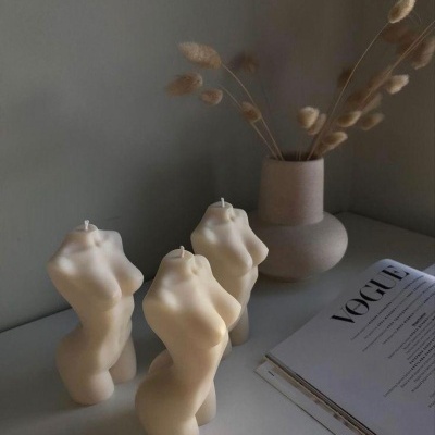 Velas em forma de torso feminino, vaso com ramos secos e revista VOGUE sobre mesa branca