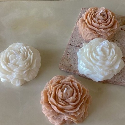 Velas em forma de flor com pétalas detalhadas, em branco e bege, sobre superfície neutra e pedra rústica