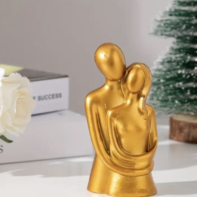 Estátua dourada de casal abraçado sobre superfície branca com flor branca e árvore de Natal