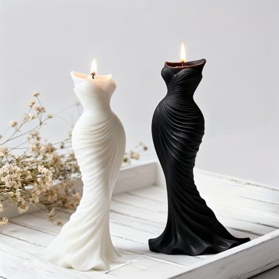 Duas velas em forma de manequins femininos vestidos, uma branca e uma preta, sobre bandeja branca com flores secas ao lado.