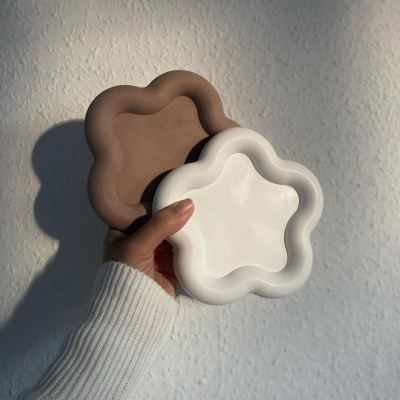 Duas bandejas decorativas em forma de flor, uma castanha e outra branca, seguradas por uma mão com camisola branca