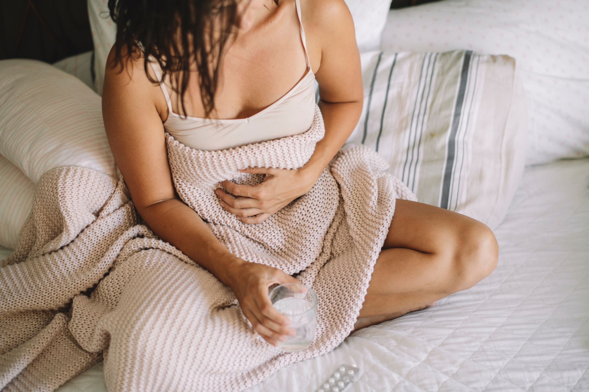 Mulher sentada na cama com cobertor bege e copo de água