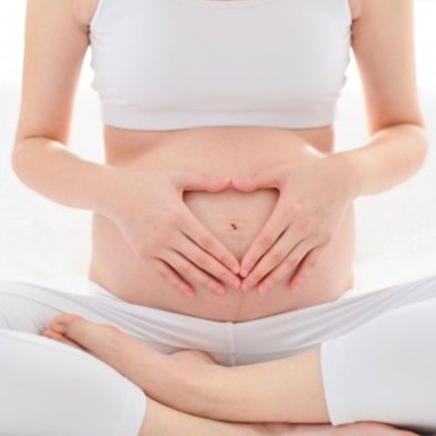 Mulher grávida sentada com roupa branca fazendo um coração com as mãos na barriga.