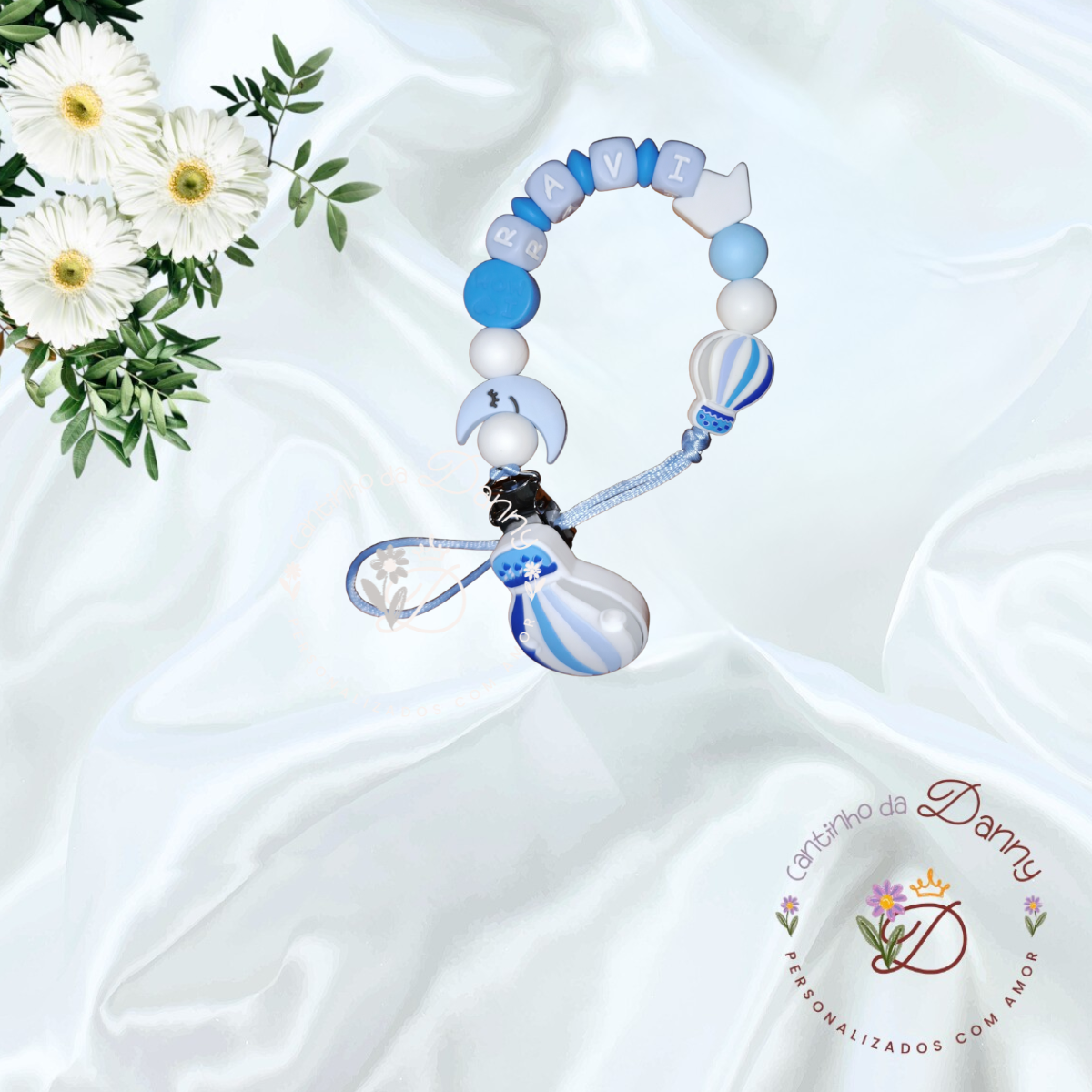 Prendedor de chupeta azul e branco com nome RAVI e pendentes, sobre tecido branco e flores brancas