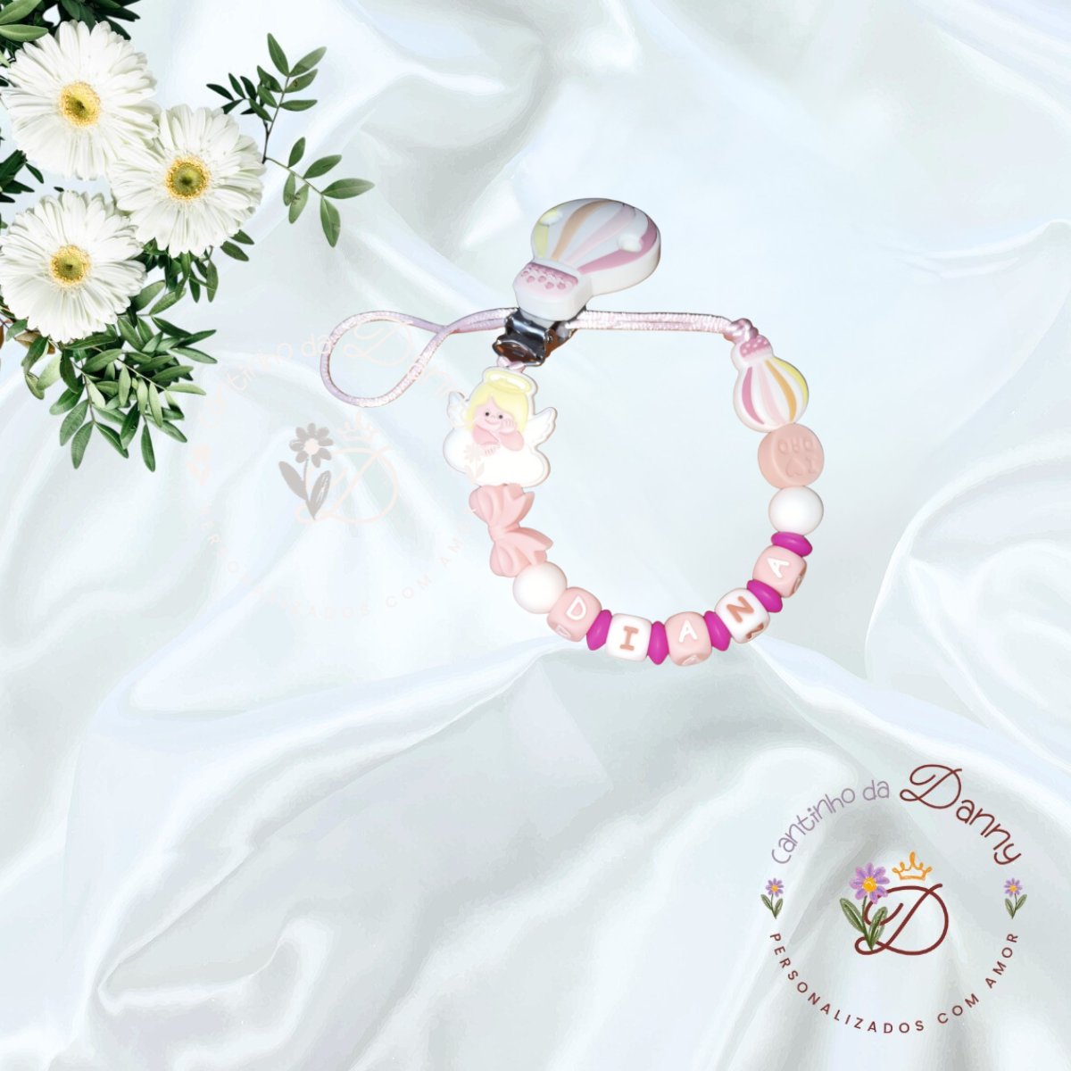 Prendedor de chupeta personalizado com nome DIANA e decoração de anjinho e balão sobre pano branco com flores