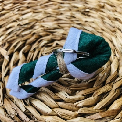 Chaveiro de tecido verde com fita branca e aro metálico sobre superfície de palha