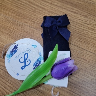 Meias de bebé azuis com laço e etiqueta 'CANTINHO DO L' sobre mesa de madeira com flor artificial roxa.