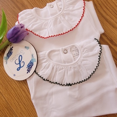 Duas blusas brancas infantis com gola rodada e folhos, bordas vermelha e preta, etiqueta 'CANTINHO L' e flor roxa sobre madeira clara