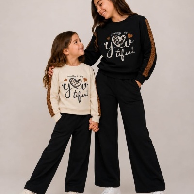 Duas raparigas com conjuntos de camisolas e calças, uma com camisola bege e outra preta, ambas com padrão animal print e texto estampado.