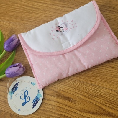 Mala de tecido rosa com estrelas brancas e bordado de personagem em aba branca sobre mesa de madeira com flores roxas e etiqueta redonda.