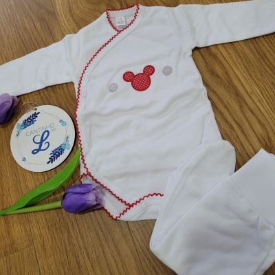 Body de bebé branco com bordado de cabeça de rato vermelho e costuras vermelhas num chão de madeira com flores roxas.