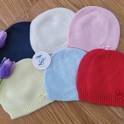 Cinco gorros de malha coloridos com laços e etiqueta AMARENO L sobre mesa de madeira com tulipas roxas.