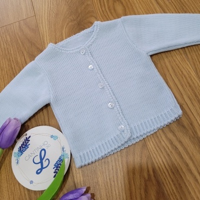 Camisola azul clara de bebé em malha com botões brancos sobre chão de madeira com flores roxas e cartão redondo