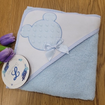 Toalha de banho azul claro para bebé com capuz branco bordado com cabeça de urso azul e laço, etiqueta redonda e flores roxas.