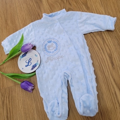 Pijama azul claro de bebé com bordado e textura bolinhas sobre chão de madeira com tulipas roxas e etiqueta redonda