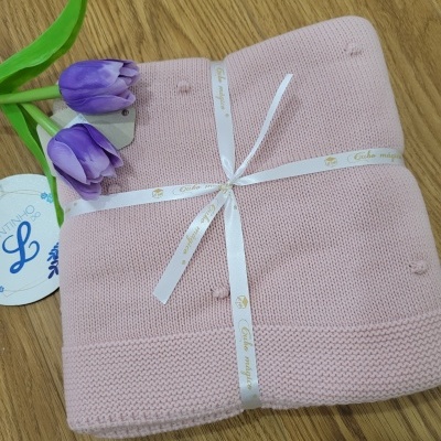 Manta de malha rosa clara para bebé, com fita branca e flores roxas, sobre madeira