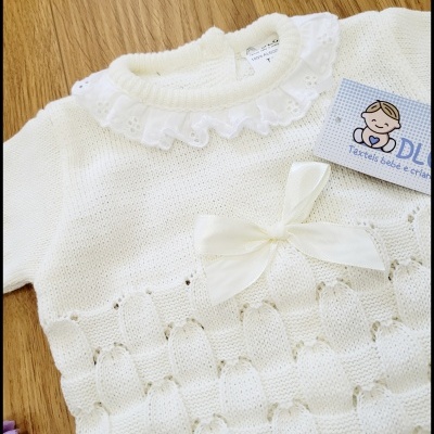 Camisola branca de malha para bebé com gola folhos e laço, sobre superfície de madeira clara.