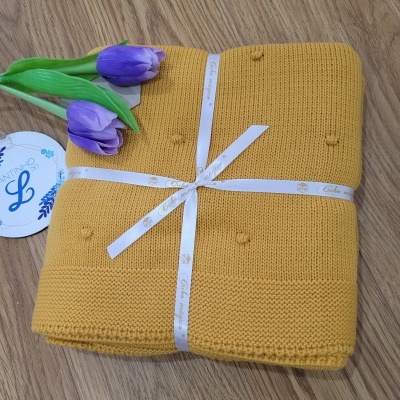 Manta amarela de malha com pompons, fita branca com texto, tulipas roxas artificiais e etiqueta branca com letra L azul