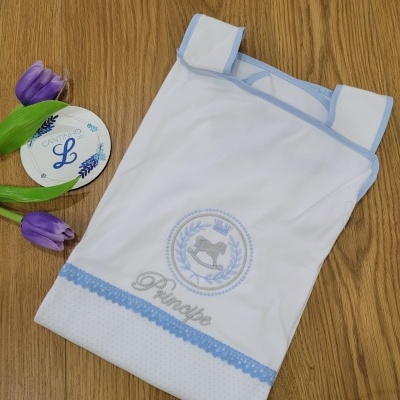 Lençol de bebé branco com bordado azul e cinza sobre madeira com tulipas roxas e etiqueta redonda